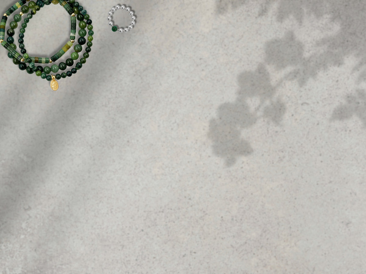 A flat-lay view of a green beaded bracelet stack with a small gold pendant and a silver beaded ring with a green gemstone, placed on a textured light gray surface with soft leaf shadows.